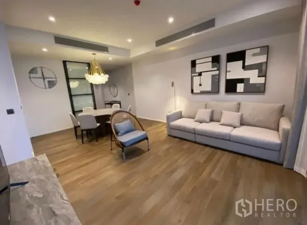 Condominium for rent in Pathum Wan, Bangkok - Spacious living–dining zone with engineered wood flooring, large sofa, modern chandelier, and glass partition door.