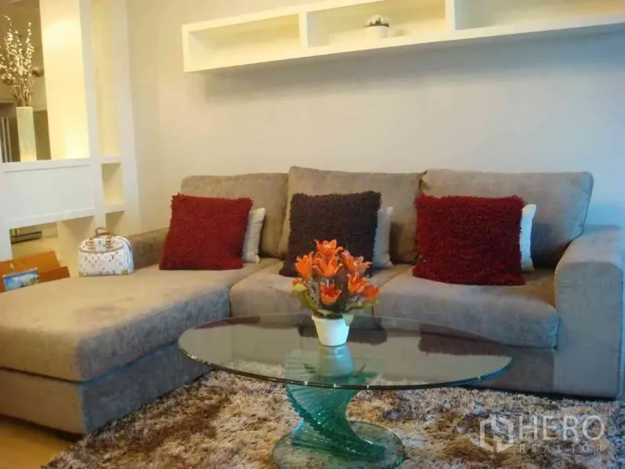 Condominium for rent in Watthana, Bangkok - Bright living room with gray L-shaped sofa, glass coffee table and floating shelves in a modern Sukhumvit condo.