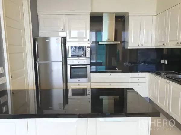 公寓 出租在Watthana, Bangkok - 现代白色厨房，黑色石材台面与中岛，配备烤箱和不锈钢冰箱等电器。