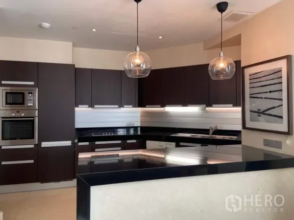 Condominium for rent in Bang Rak, Bangkok - Sleek U-shaped kitchen with dark cabinetry, stone countertops, stainless appliances, and two glass pendant lights.