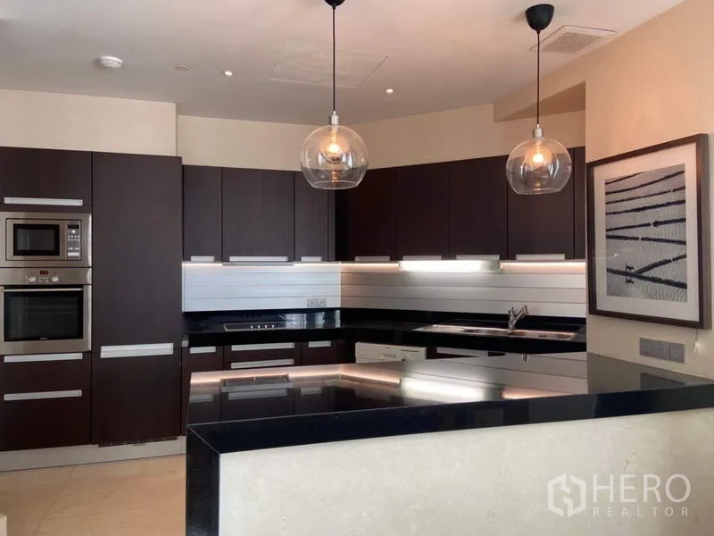 Condominium for rent in Bang Rak, Bangkok - Sleek U-shaped kitchen with dark cabinetry, stone countertops, stainless appliances, and two glass pendant lights.
