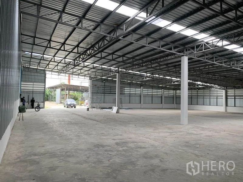 货栈 出租在Mueang Pathum Thani, Pathum Thani - 仓库内部宽敞空间，高钢结构屋顶与采光板，混凝土地面，后方为车辆出入口。