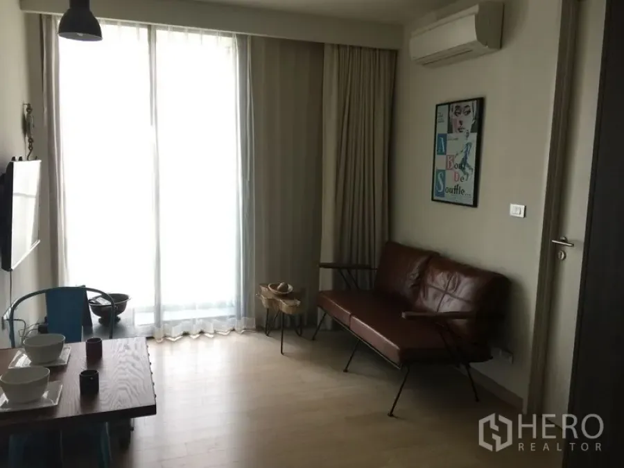 公寓 出租在Watthana, Bangkok - 明亮客厅，棕色皮沙发、落地窗帘、壁挂空调与用餐角。