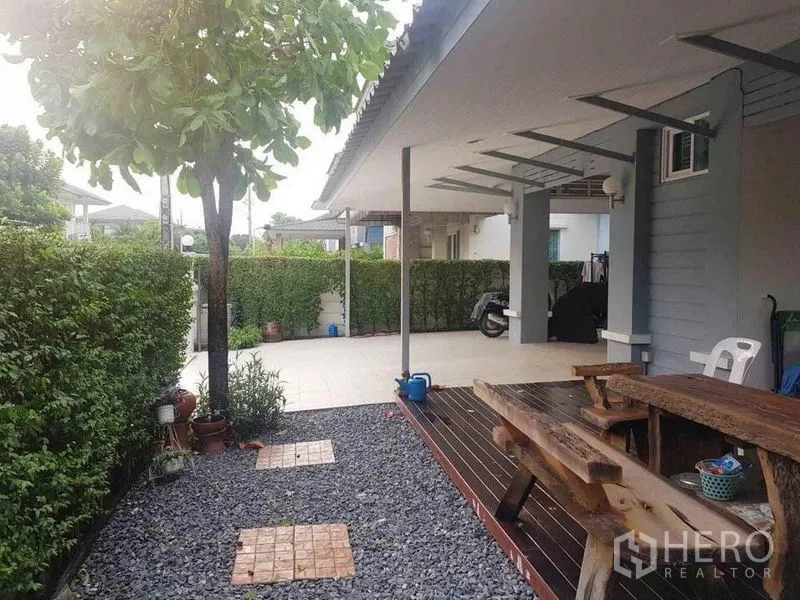 Detached House for sale in Nonthaburi - Side yard with trimmed hedges, wooden deck table and pebble garden.