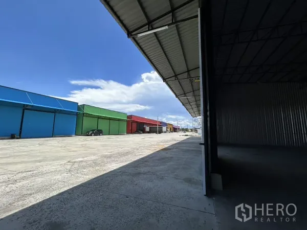 Warehouse for rent in Ban Pho, Chachoengsao - Covered loading apron facing a spacious concrete yard with colorful neighboring warehouse units and parking area.
