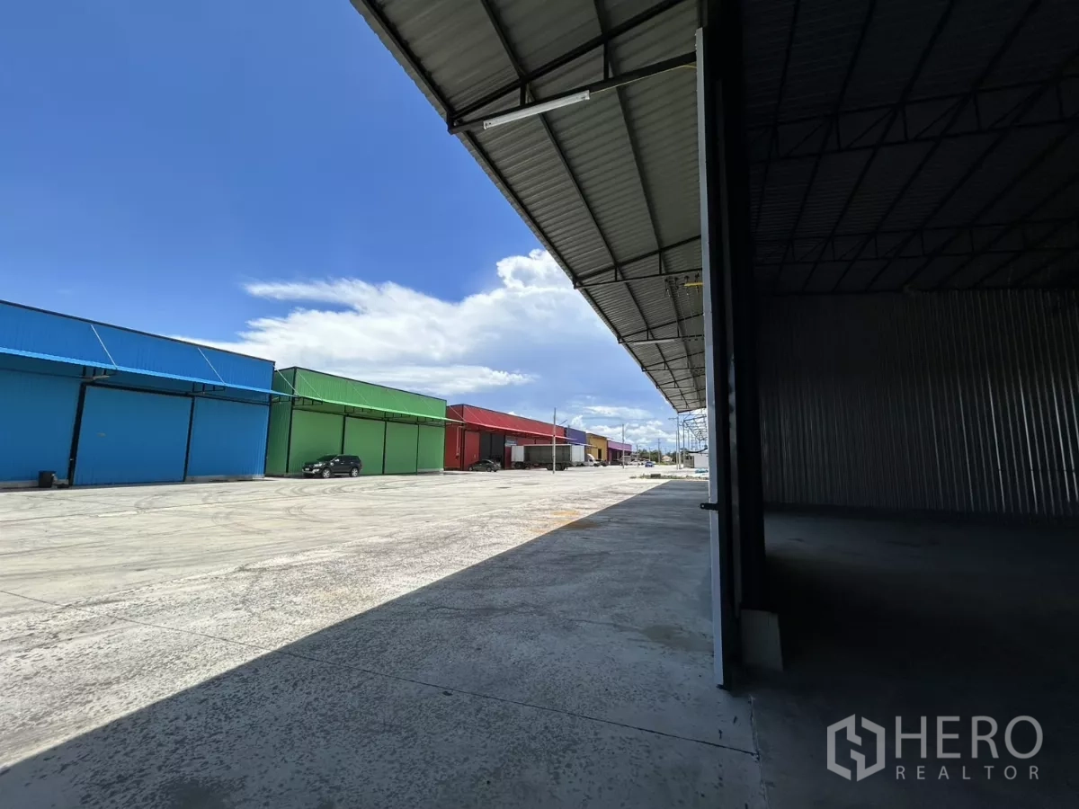 Warehouse for rent in Ban Pho, Chachoengsao - Covered loading apron facing a spacious concrete yard with colorful neighboring warehouse units and parking area.