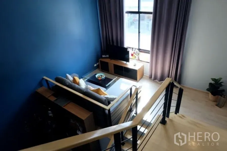 Townhouse for rent in Khlong Toei, Bangkok - Staircase landing overlooking the living area with TV console, large window and grey sofa.