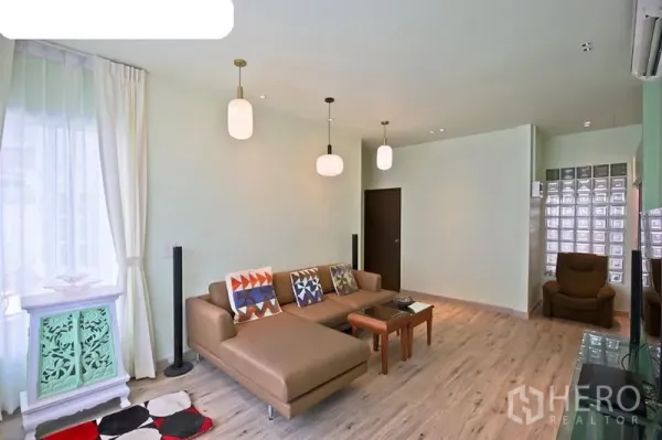 Detached House for rent in Bang Kapi, Bangkok - Open-plan lounge with tan sectional sofa, pendant lighting, wood floors, and a glass-block accent wall.