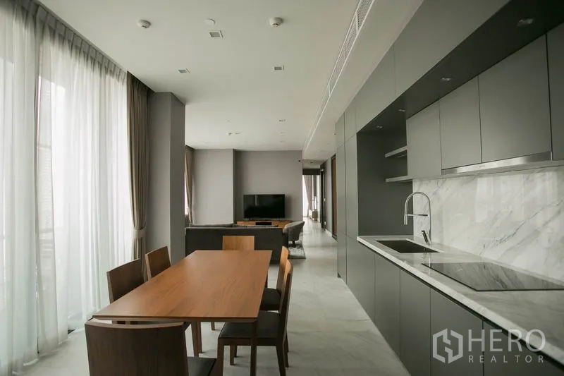 Condominium for rent or sale in Watthana, Bangkok - Open-plan dining and sleek kitchen with marble backsplash, long counter, built-in hob and wood dining table.