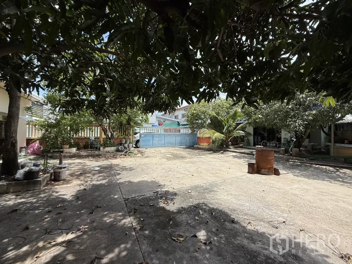 Warehouse for sale in Phra Samut Chedi, Samut Prakan - Large open courtyard shaded by trees facing a blue metal entrance gate.