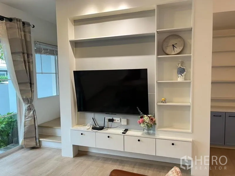 联排别墅 出租在Prawet, Bangkok - 客厅带内置电视墙与开放式搁架，浅色木地板与平板电视。