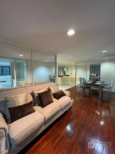 Condominium for rent - Open-plan living and dining area with polished wood floors, beige sofa, glass dining table, mirrored wall, and recessed lighting.