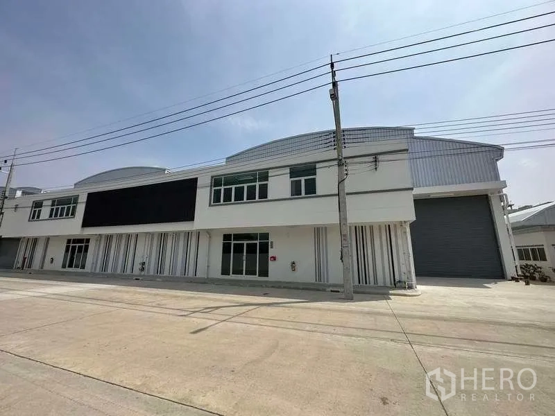 Factory for rent in Khlong Luang, Pathum Thani - Modern two-storey factory facade with large roller shutter and wide concrete frontage under clear sky.