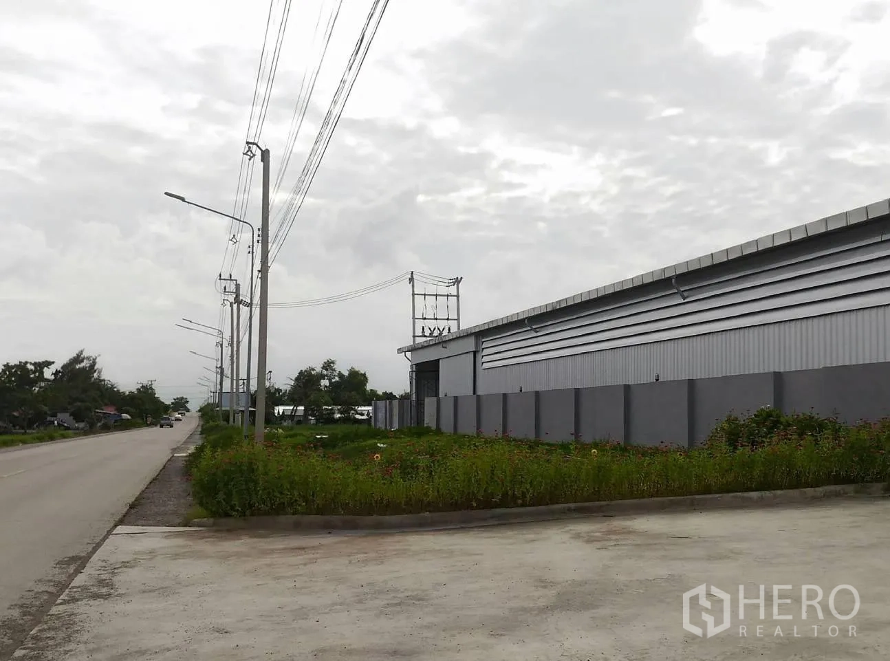 货栈 出租在Lam Luk Ka, Pathum Thani - 临路仓库外观，灰色金属墙体与围墙，电线杆成排，宽阔混凝土出入口