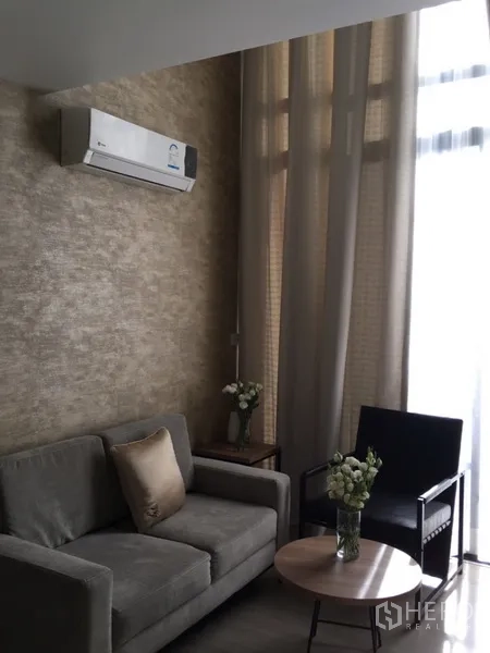 Condominium for sale in Pathum Wan, Bangkok - Living area featuring a sofa, armchair, round coffee table, air conditioner, and tall windows with sheer drapes.