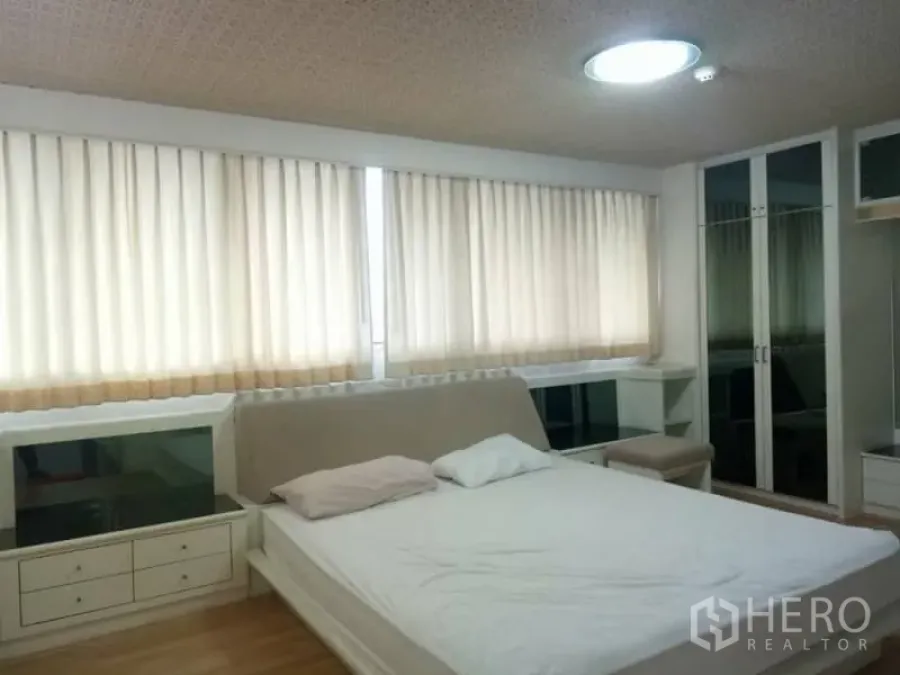 Condominium for rent in Watthana, Bangkok - Bright master bedroom with large windows, beige curtains, built-in storage and a mirrored wardrobe over wood flooring.