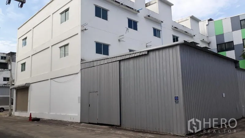 อาคารพาณิชย์ ให้เช่าในLam Luk Ka, Pathum Thani - ภายนอกอาคารพาณิชย์หลายชั้นสีขาว พร้อมโกดังเมทัลชีทและทางเข้าในซอย