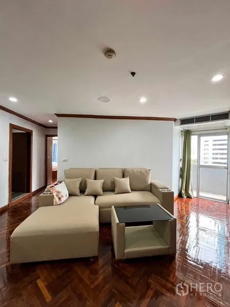 Condominium for rent in Ratchathewi, Bangkok - Bright living room with a beige sectional sofa, glass coffee table, balcony door and polished parquet floors.