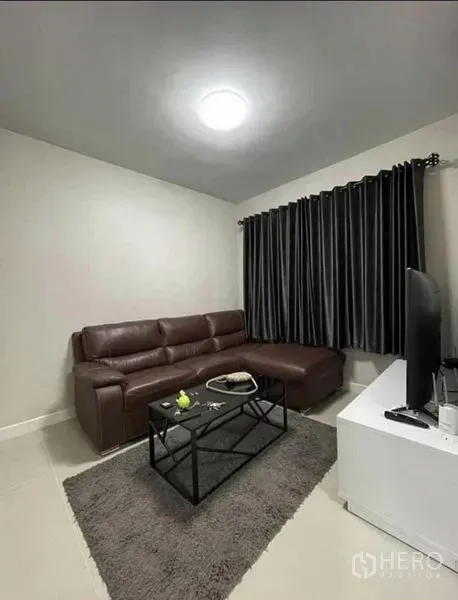 สำนักงานที่บ้าน ให้เช่าในSamut Prakan - มุมนั่งเล่นอบอุ่น โซฟาหนัง พรม โต๊ะกลาง และทีวีบนตู้สีขาว พร้อมผ้าม่านทึบ