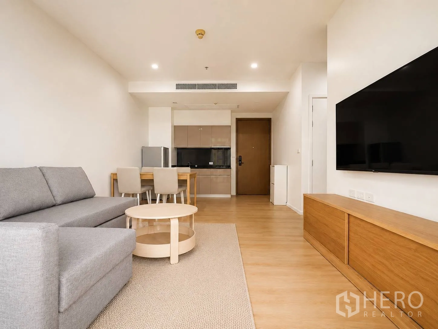 Condominium for rent or sale in Watthana, Bangkok - Open-plan living room with grey sofa, round coffee table, dining set, kitchenette and wall-mounted TV on wood floors.