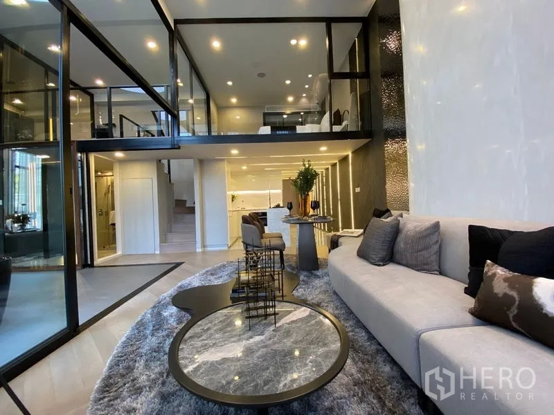 Detached House for sale in Watthana, Bangkok - Wide living zone with large sofa, nested coffee tables, and a glass-encased mezzanine.