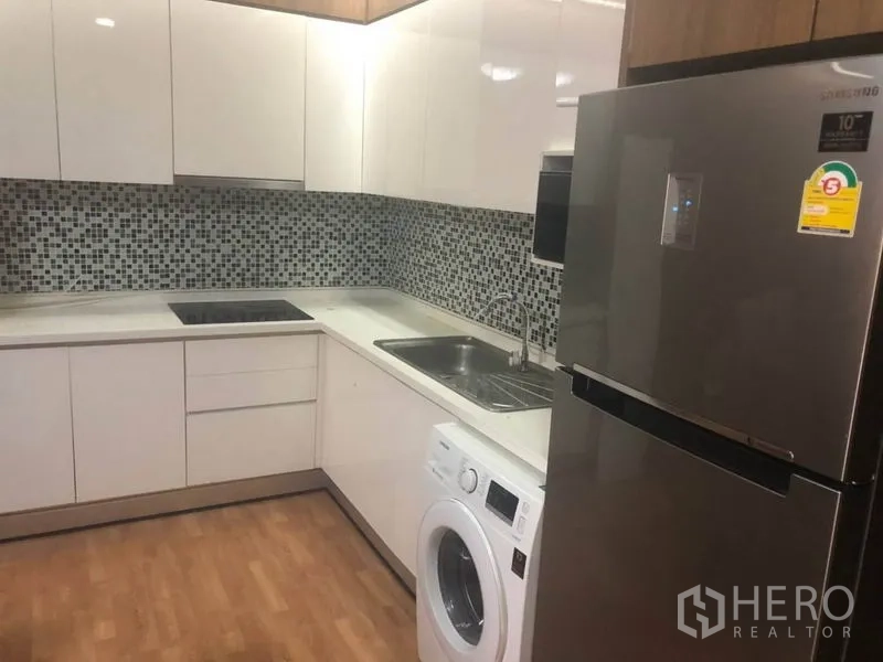 Condominium for sale in Bang Na, Bangkok - Modern U-shaped kitchen with glossy white cabinets, mosaic tile backsplash, fridge, cooktop and washing machine.