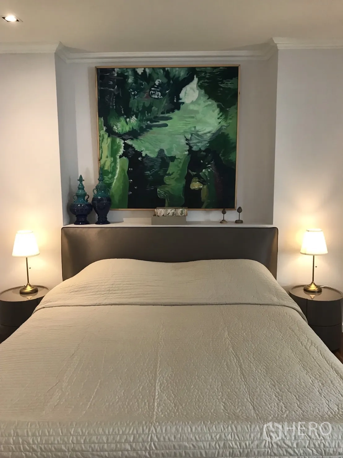 คอนโด ให้เช่าในBangkok - ห้องนอนอบอุ่น เตียงหัวเตียงบุผ้า โคมไฟข้างเตียง และภาพศิลปะสีเขียว