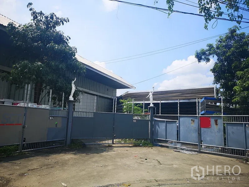 货栈 出租在Bang Khen, Bangkok - 曼谷仓库入口的金属滑动大门，周围绿树成荫。