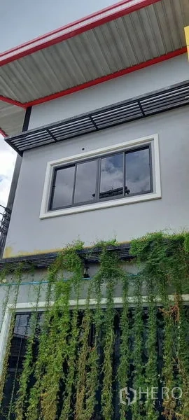 Warehouse for rent or sale in Krathum Baen, Samut Sakhon - Modern warehouse facade with gray walls, black-framed window, canopy and trailing green vines.