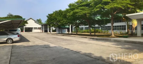 工厂 出售在Mueang Samut Sakhon, Samut Sakhon - 园区内部宽阔车道与遮阴停车区，旁有树木与厂房立面。