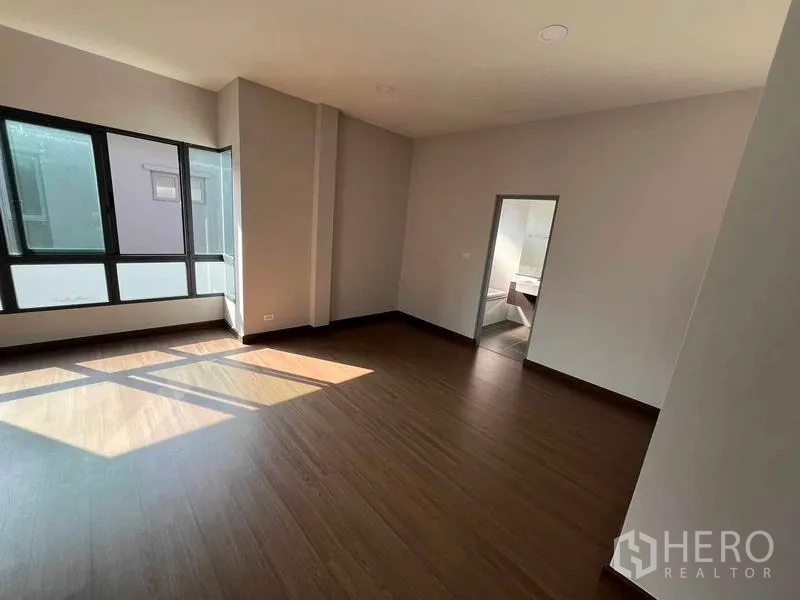 Detached House for rent in Khlong Sam Wa, Bangkok - Sunlit bedroom with wood floor, large windows and doorway to an ensuite bathroom.