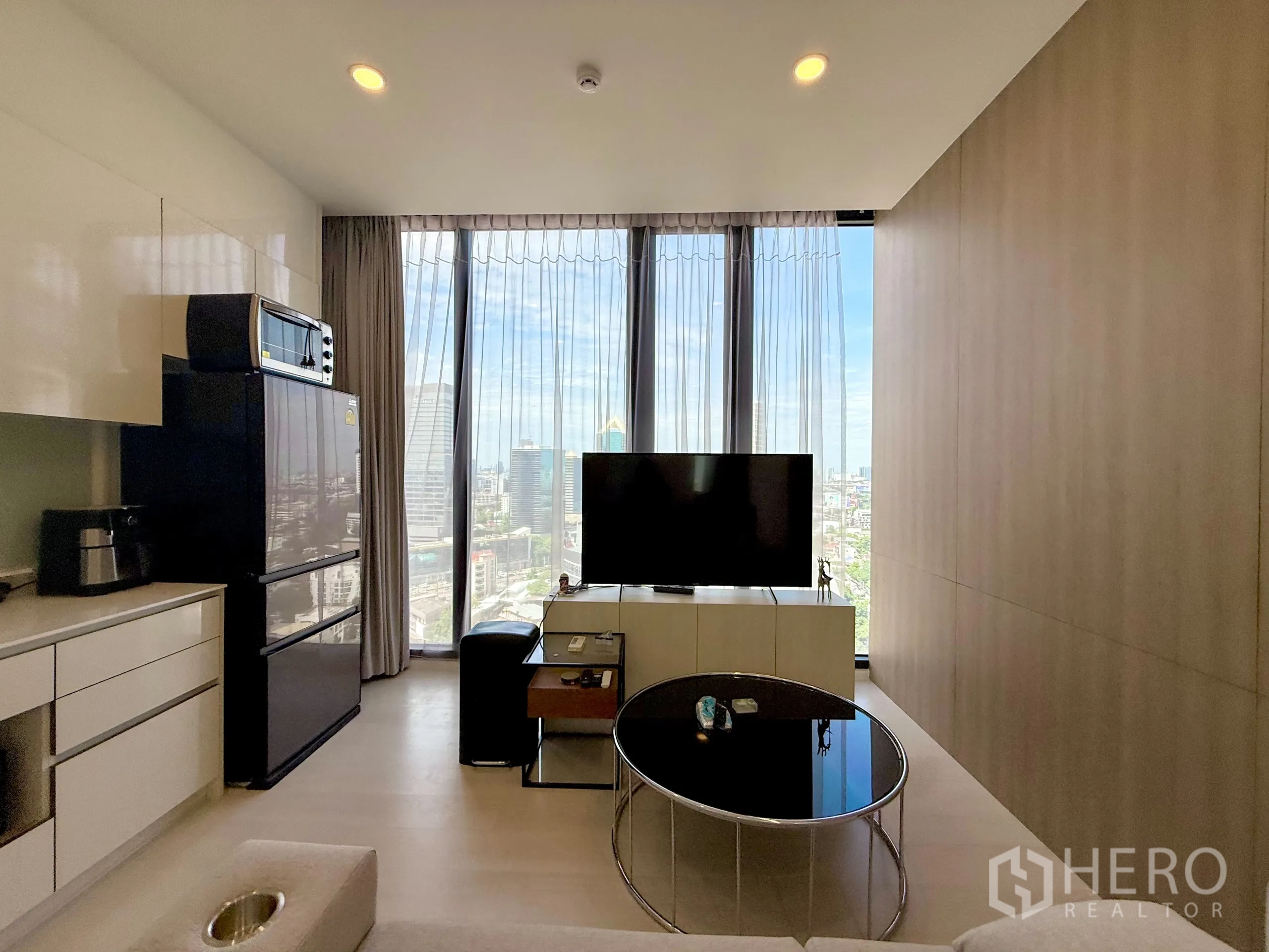 Condominium for sale in Chatuchak, Bangkok - Living room with TV and open kitchen facing tall glass windows with city view.