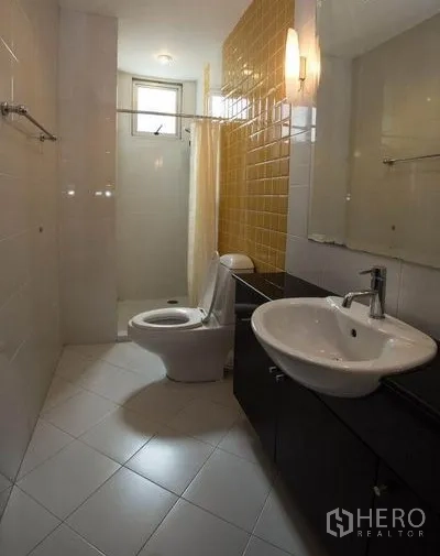 Condominium for rent in Khlong Toei, Bangkok - Narrow bathroom with yellow accent tiles, toilet, countertop basin, and shower area.