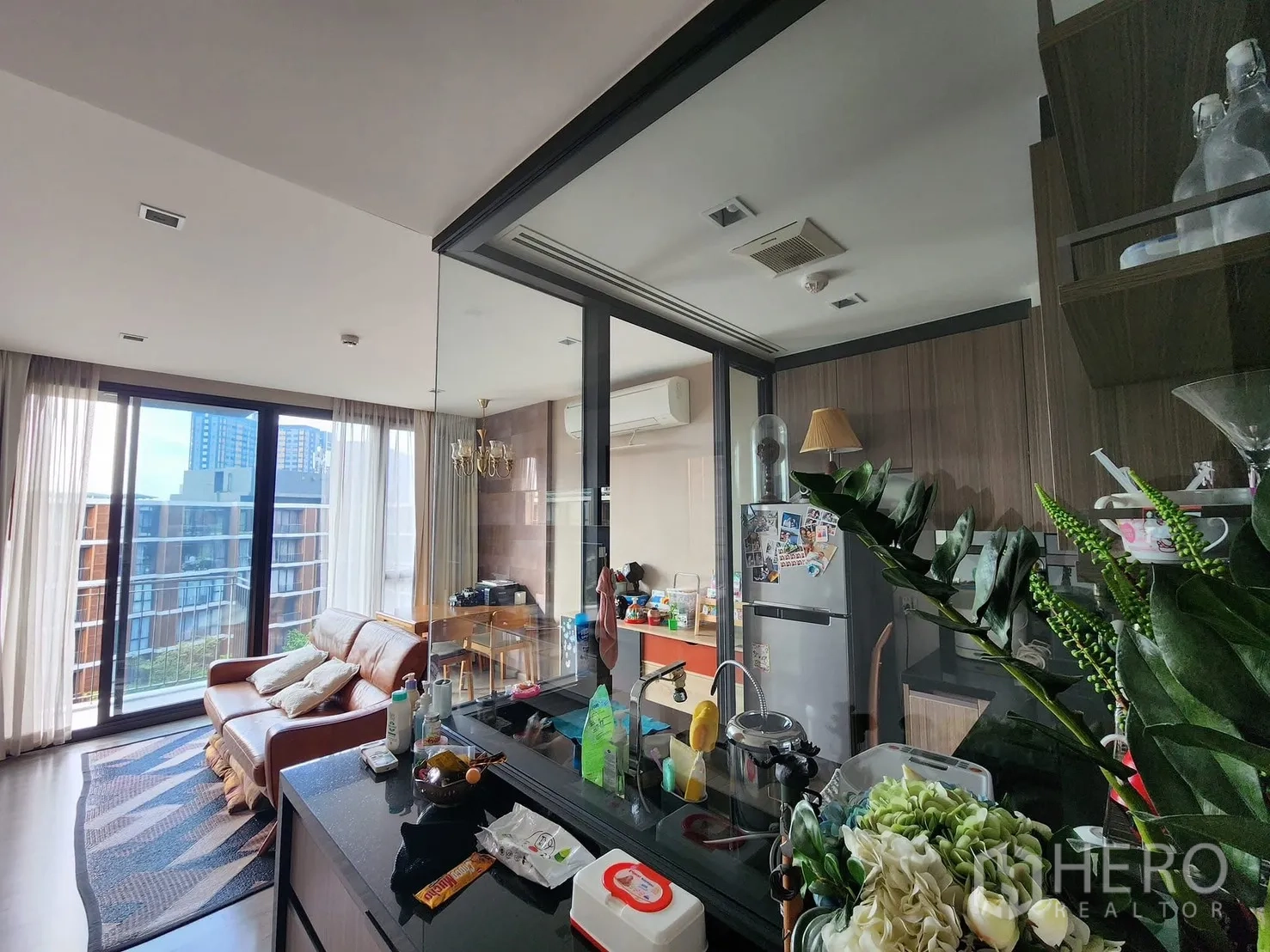 Condominium for rent in Watthana, Bangkok - Bright living room with floor-to-ceiling windows, leather sofa, and a glass-partitioned kitchen with wood cabinetry.