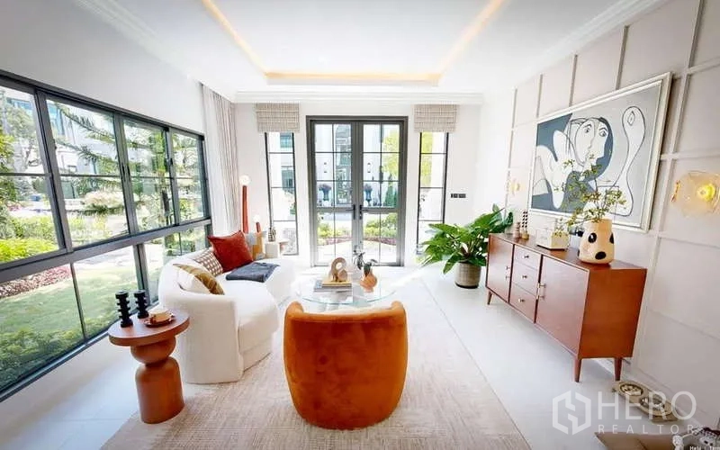 Detached House for rent in Bang Phli, Samut Prakan - Cozy living room with large black-framed windows, modern art and mid-century wood furniture overlooking landscaped greenery.