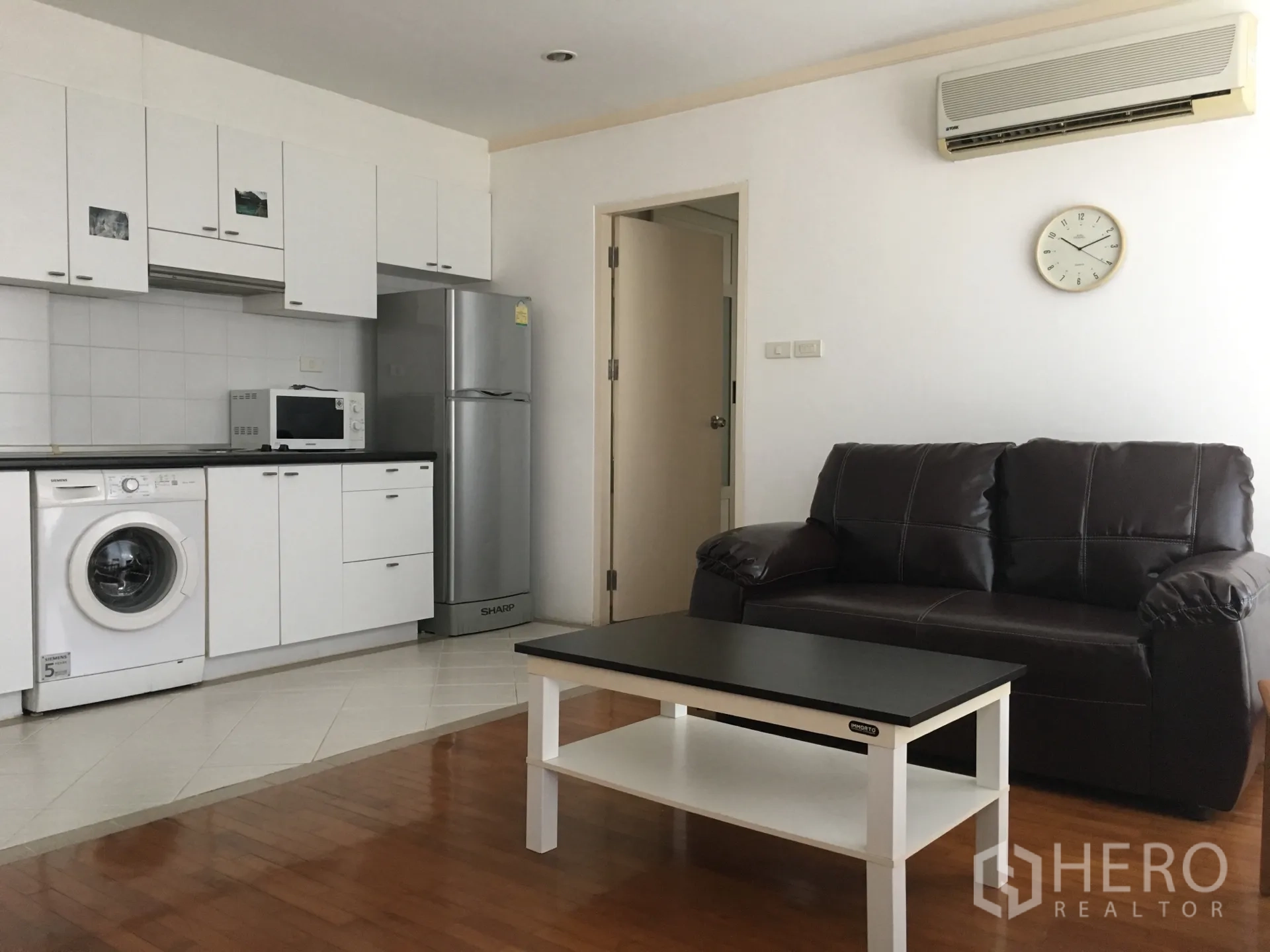 Condominium for rent or sale in Watthana, Bangkok - Bright living area with leather sofa, coffee table, wall AC and a white kitchenette with washer, microwave and fridge.