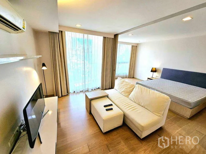 Condominium for rent - Sunny living space with sofa, TV and bed by large curtained windows.