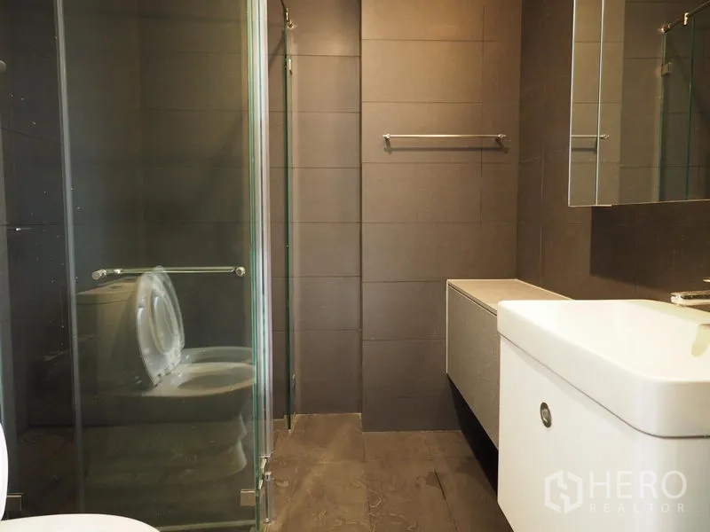 Condominium for rent in Pathum Wan, Bangkok - Contemporary bathroom with glass-enclosed shower, wall-mounted vanity and dark tiles.