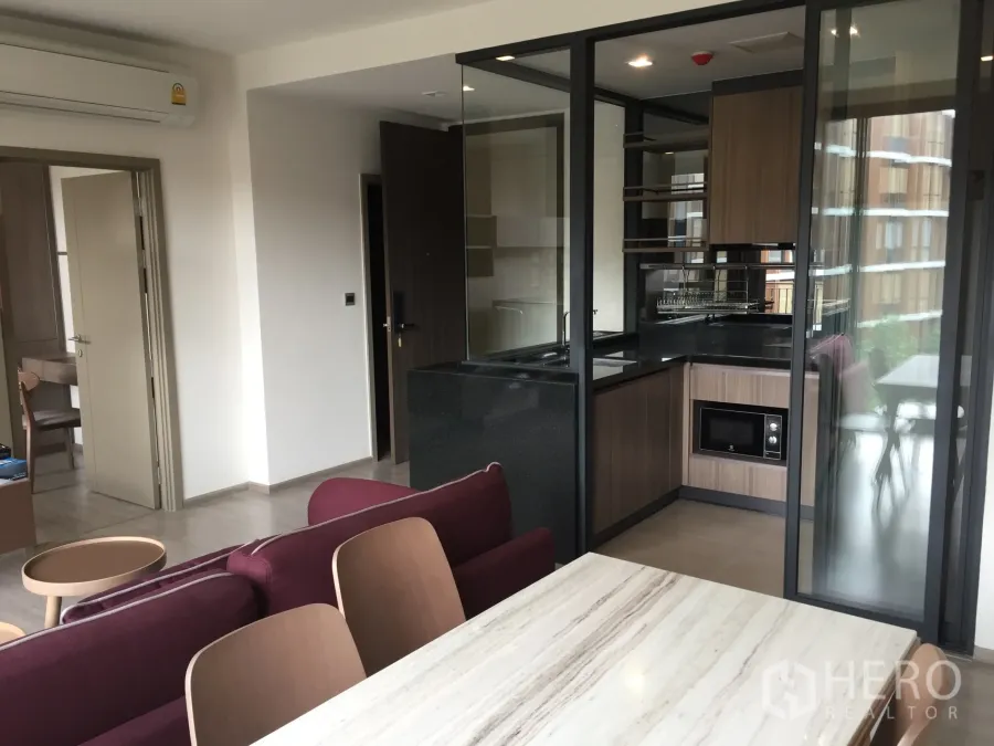 Condominium for rent in Watthana, Bangkok - Open-plan living with marble dining table and a glass-partitioned L-shaped kitchen with wood cabinetry and built-in microwave.