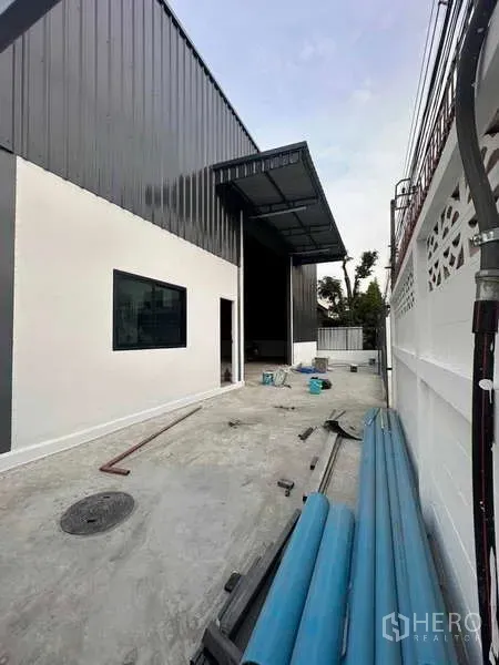 货栈 出租在Bang Khen, Bangkok - 仓库侧院，灰色外立面带雨棚，宽阔混凝土通道放置管材。