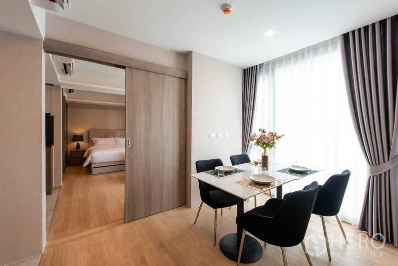 Condominium for rent in Huai Khwang, Bangkok - Dining area with four chairs by a large window and sliding door to the bedroom.