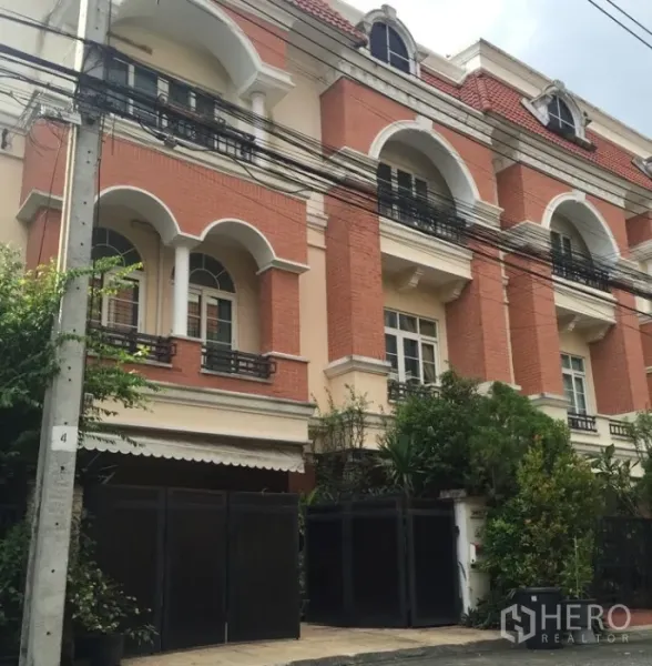 ทาวน์เฮาส์ ให้เช่าหรือขายในBueng Kum, Bangkok - ด้านหน้าทาวน์เฮาส์สามชั้น ผนังอิฐแดง ซุ้มโค้ง ระเบียง และรั้วสีเข้มรายล้อมด้วยต้นไม้
