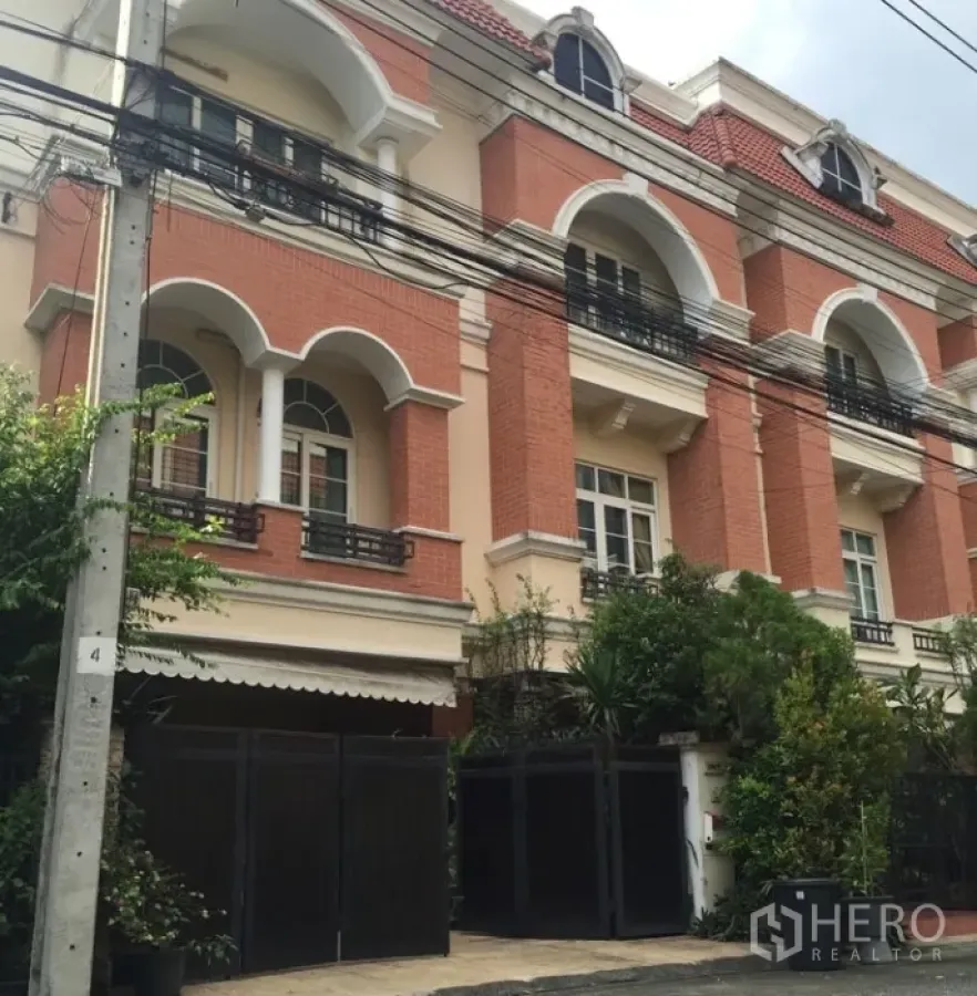 Townhouse for rent or sale in Bueng Kum, Bangkok - Three-storey townhouse exterior with red-brick facade, arched windows, balconies and a gated entrance framed by greenery.