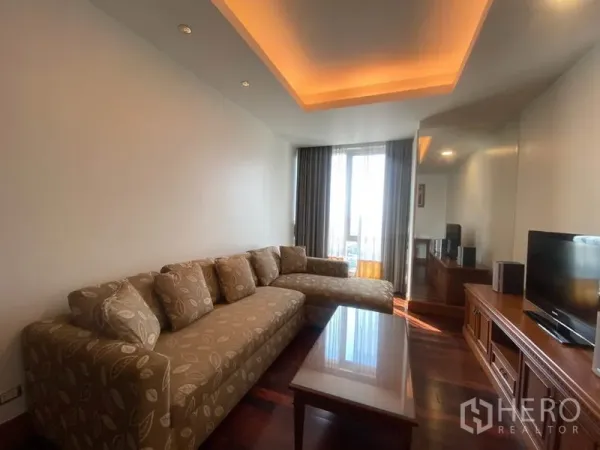 Condominium for rent in Sathon, Bangkok - Cozy living room with patterned L-shaped sofa, cove lighting, coffee table, wood floors and tall window.