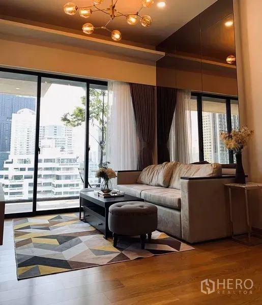 Condominium for rent in Watthana, Bangkok - Warm living room with sofa, geometric rug and floor-to-ceiling glass opening to a balcony with city views.