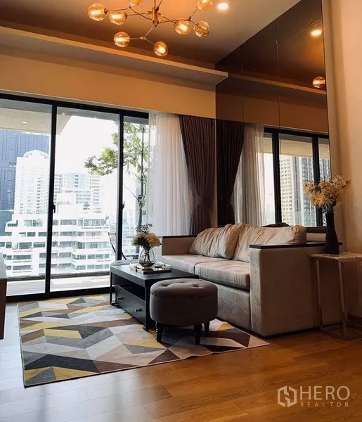 Condominium for rent in Watthana, Bangkok - Warm living room with sofa, geometric rug and floor-to-ceiling glass opening to a balcony with city views.