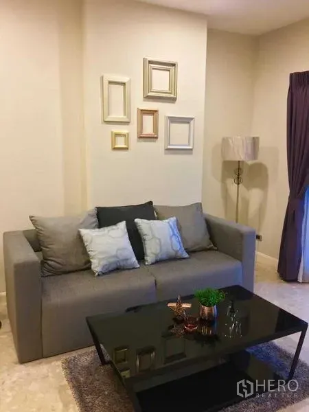 คอนโด ให้เช่าในKhlong Toei, Bangkok - ห้องนั่งเล่นโทนอบอุ่น โซฟาสีเทา หมอนอิง โต๊ะกลางกระจก ผนังสีอ่อนและโคมไฟมุมห้อง