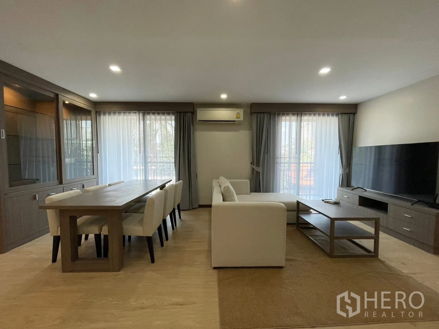 Condominium for rent in Watthana, Bangkok - Dining area for eight beside a white sofa and TV unit, framed by two wide windows with sheer curtains.