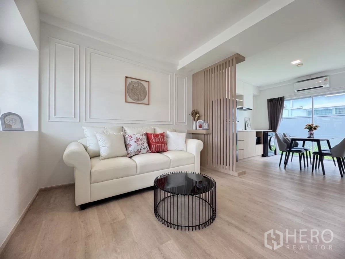 联排别墅 出租在Prawet, Bangkok - 明亮客厅，白沙发与木质格栅隔断，连接餐区与窗户。