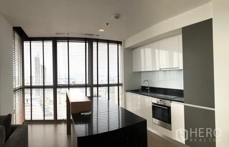 Condominium for rent or sale in Khlong San, Bangkok - Contemporary kitchen with island, white cabinets, built-in oven and expansive window blinds.